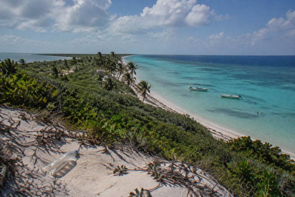 Wide view of a low sandy island with palms and boats anchored offshore under scattered clouds