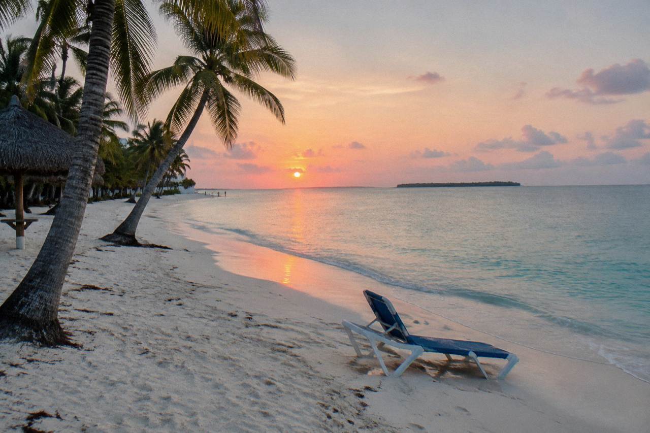 Quiet hotel beach at sunrise looking north across calm turquoise water toward a distant low island